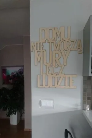 Napis: 'Domu nie tworzą mury, lecz ludzie' wycięty laserem z naturalnej sklejki, zamocowany na ścianie