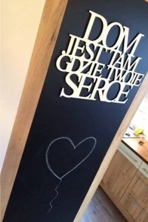 Napis: 'Dom jest tam, gdzie twoje serce' wycięty laserem z jasnej sklejki, zamocowany na tablicy kredowej
