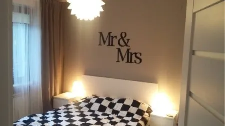 Napisy Mr & Mrs na ścianie nad łóżkiem w nowoczesnej sypialni, z lampkami nocnymi i geometryczną pościelą.