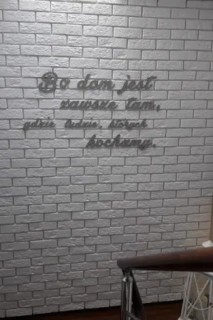 Napis: 'Bo dom jest zawsze tam, gdzie ludzie, których kochamy' wycięty laserem z naturalnej sklejki, zamocowany na białej ceglanej ścianie