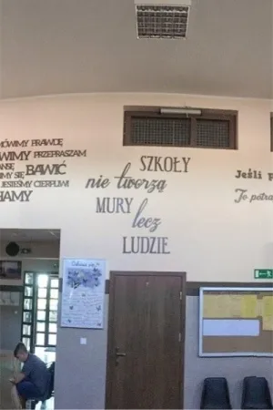Napis: 'Szkoły nie tworzą mury, lecz ludzie' wycięty laserem z ciemnej sklejki, zamocowany na ścianie w przestrzeni szkolnej